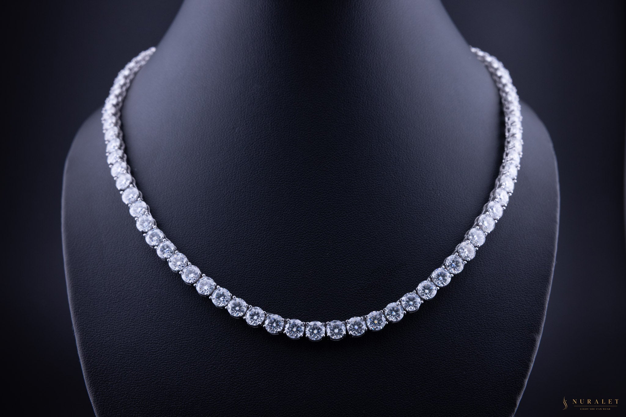 A sparkling diamond tennis necklace displayed on a black jewelry bust against a dark background.